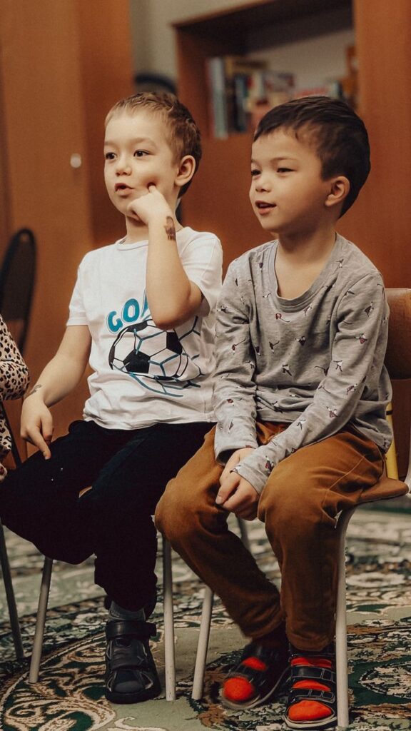 Two young boys sitting indoors, engaged in conversation, wearing casual clothes.